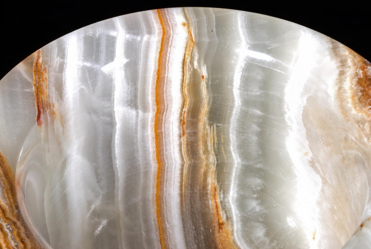 A polished, curved slice of banded onyx stone with white and grey layers and thin orange-brown stripes against a black background.