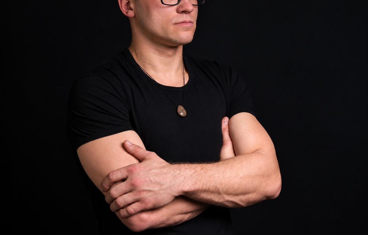 A man in a black t-shirt wears a pendant with an oval, brownish stone on a thin cord against a dark background.