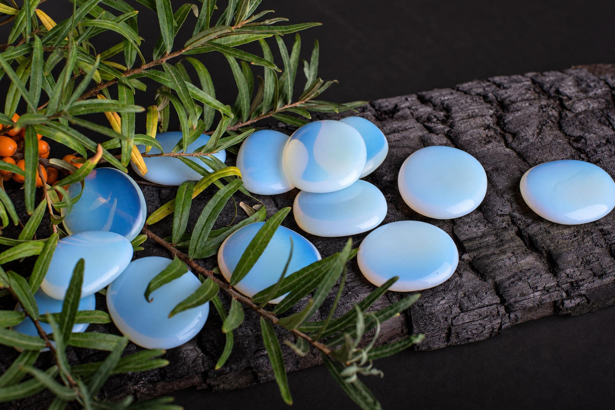 Several smooth, round blue and white opal palmstones rest on a dark wooden surface next to green leaves.