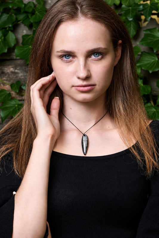 A woman wears a black top and a dark, elongated pendant with light streaks on a thin cord against green leaves.