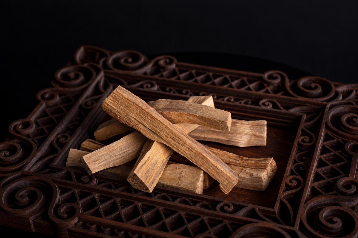 Several light brown Palo Santo wood sticks rest on a dark, ornately carved wooden tray against a black background.