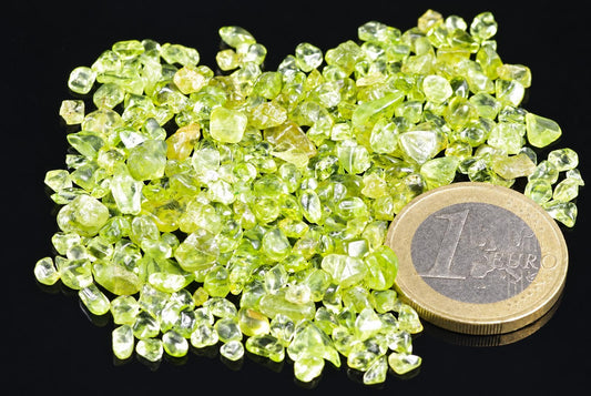 A pile of small, rough-cut green gemstones sits on a dark surface next to a one euro coin.