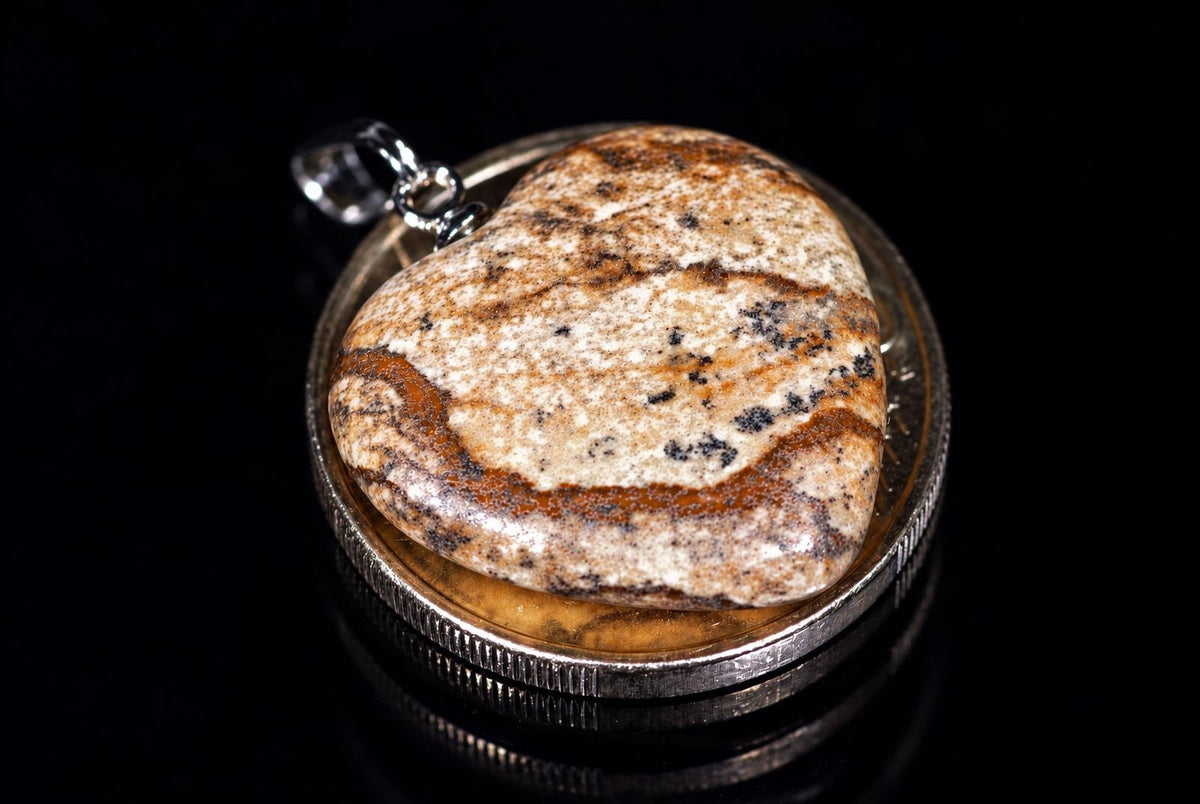 A heart-shaped jasper pendant with brown, black and white speckles sits in a silver-toned bezel on a dark surface.