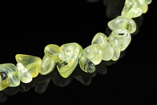 A strand of translucent yellow-green prehnite chips with dark inclusions, arranged on a dark reflective surface.