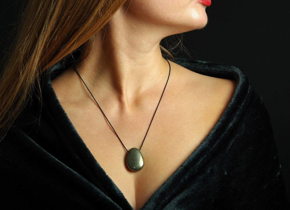 Woman wearing a natural polished Pyrite pendant – Drop on a thin black cord necklace against a dark textured fabric background, highlighting the mineral's metallic luster.