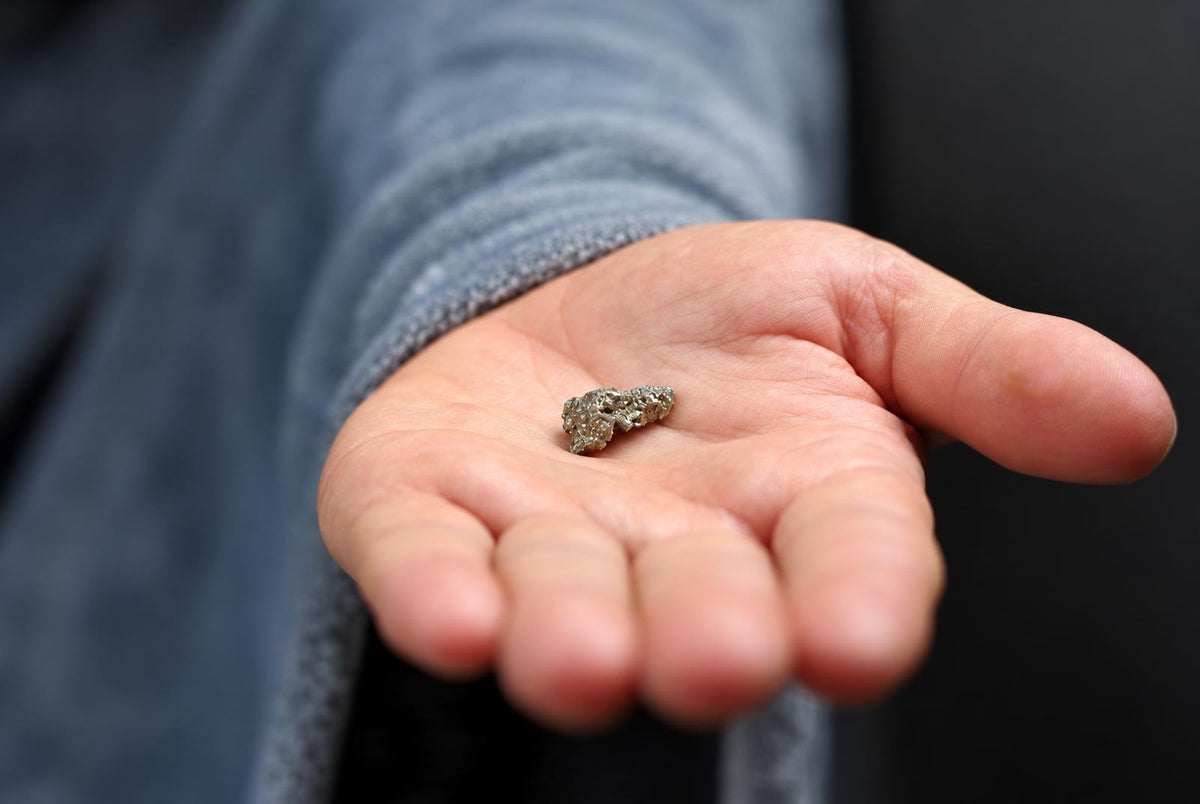 A small, rough, silver-toned mineral rests in an open palm against a dark background.