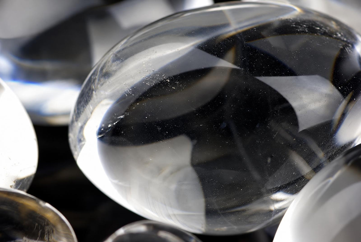 A close-up of a clear, smooth quartz crystal resting on a dark surface with other crystals nearby.