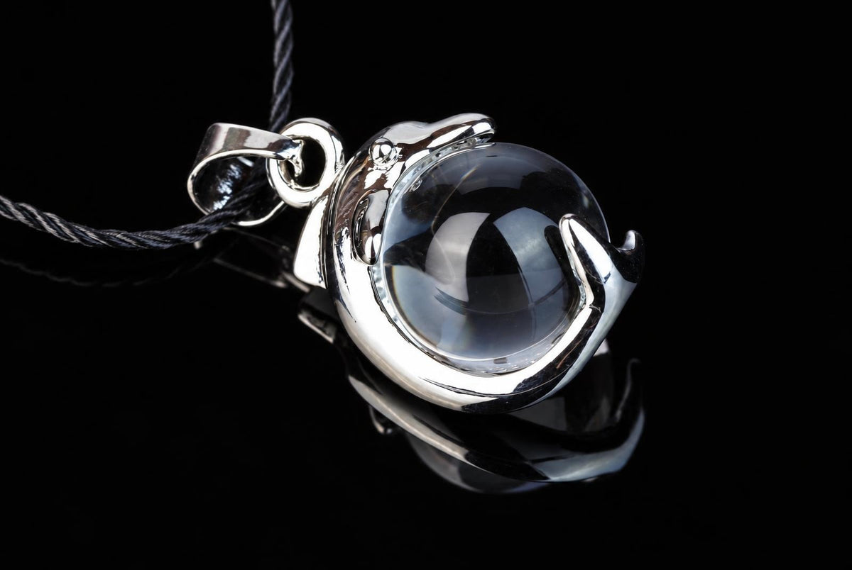 A silver-toned dolphin-shaped pendant holds a clear quartz sphere, suspended on a dark cord against a black reflective surface.