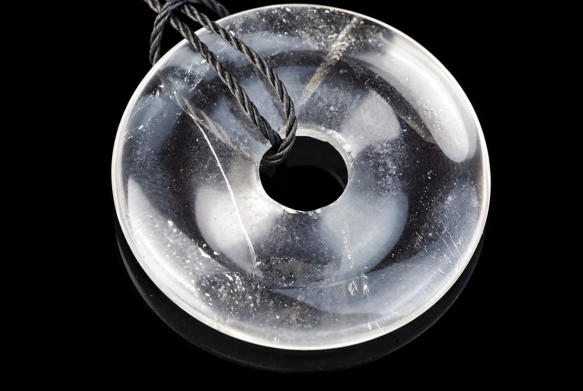 A clear quartz pendant with white swirls, a central hole, and a black cord looped through it against a dark surface.