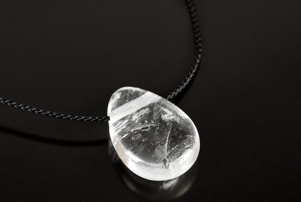 A clear quartz pendant with internal inclusions hangs from a black cord on a dark surface.