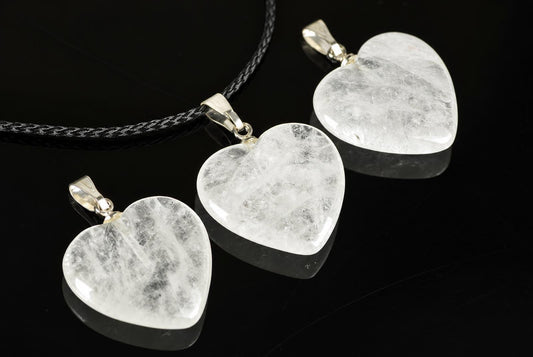 Clear quartz heart pendants with cloudy internal markings lie on a reflective black surface, with one attached to a black cord.