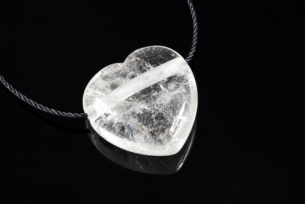 A clear, heart-shaped quartz pendant with internal white inclusions hangs from a dark cord against a black surface.