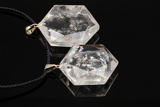 Two clear hexagonal quartz pendants with visible internal textures and gold-colored loops rest on black cords against a dark background.