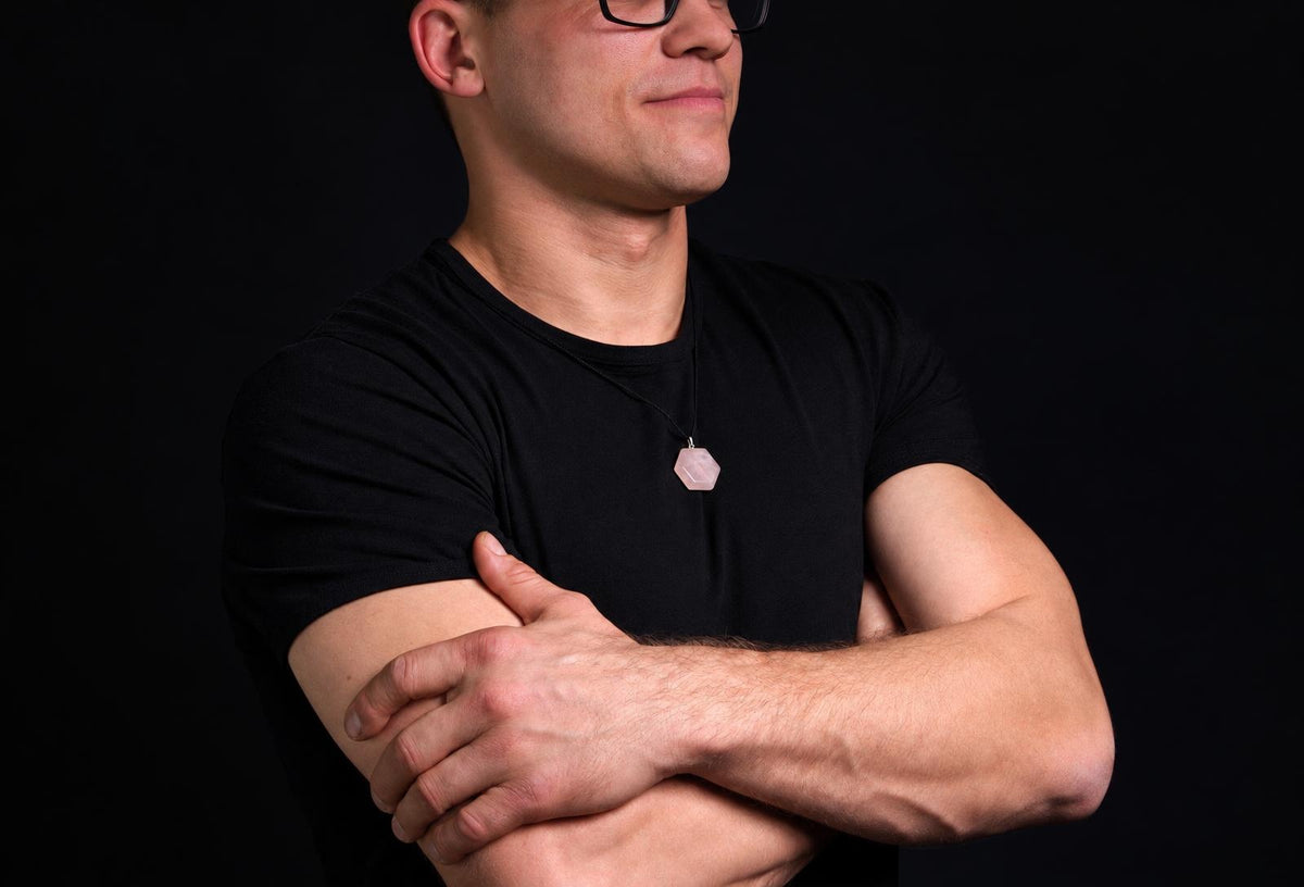 A man in a black t-shirt wears a necklace with a pale pink hexagonal pendant on a dark surface.