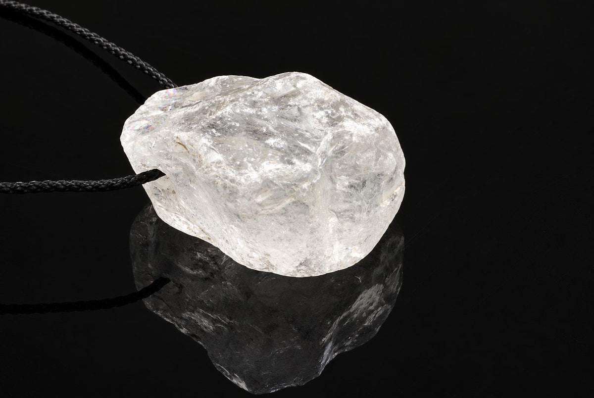 A clear, rough quartz crystal pendant on a dark cord rests on a dark surface with a reflection.