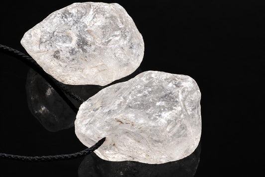 Two clear quartz stones on black cords, resting on a dark surface against a black background.
