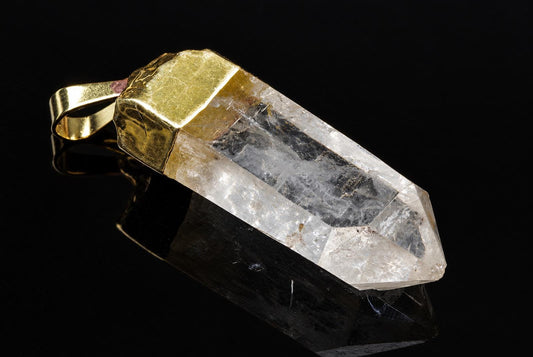 A clear pointed quartz pendant with cloudy internal textures and a gold-colored cap lies diagonally against a reflective black background.