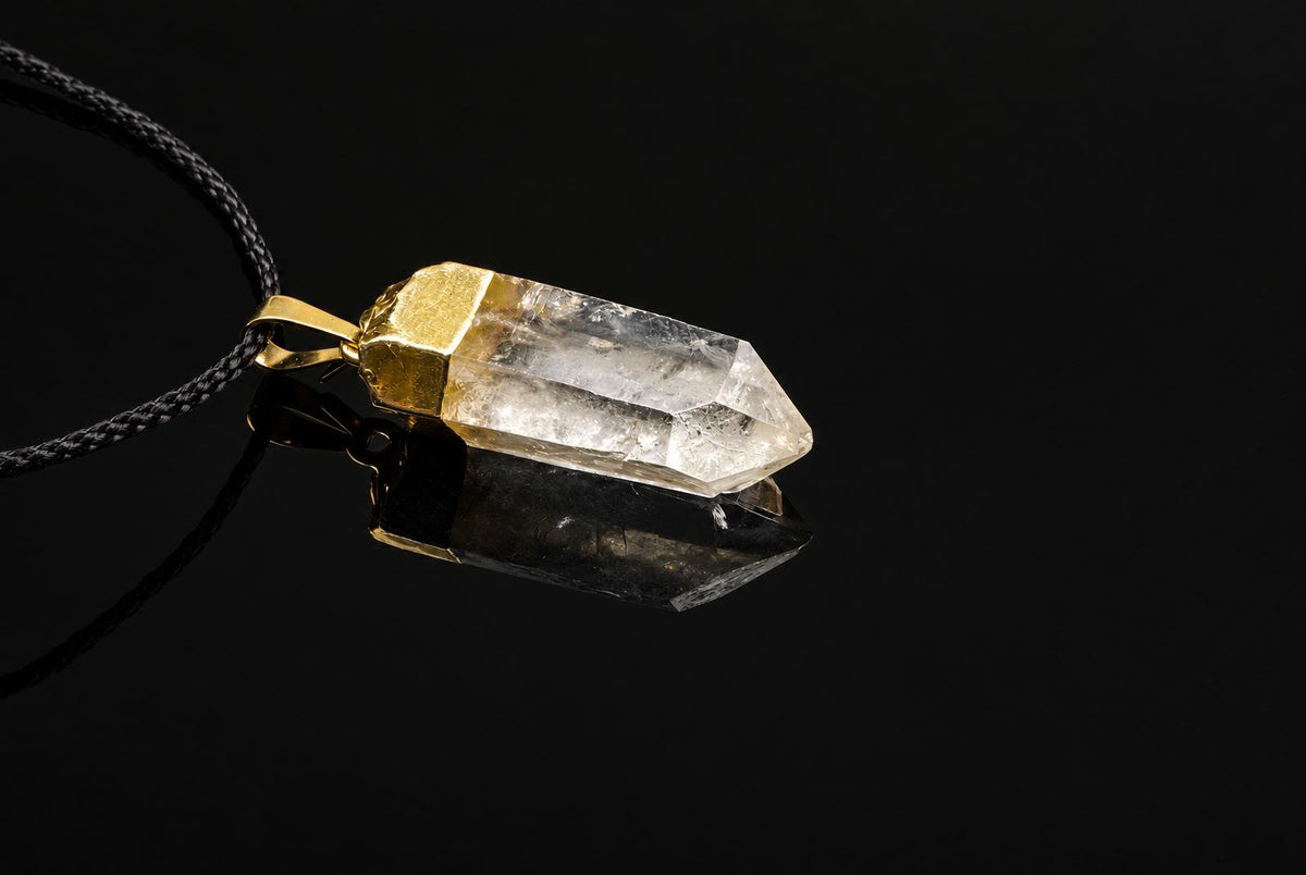 A quartz crystal pendant with a gold-coloured cap hangs from a dark cord on a black reflective surface.