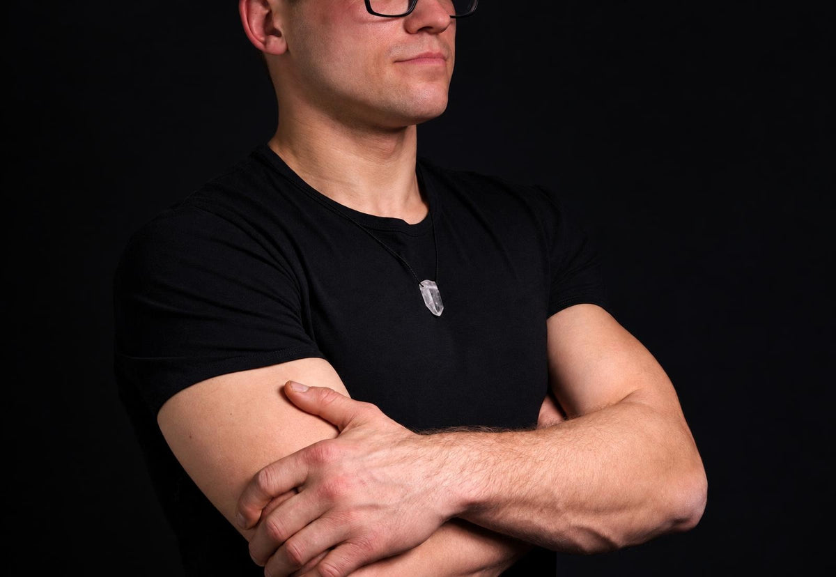 A man in a black t-shirt wears a necklace with a clear quartz pendant on a dark surface.