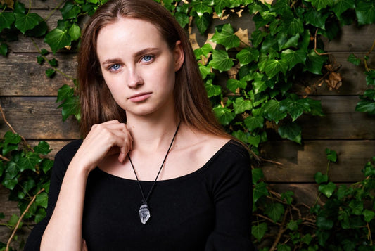 A woman wears a black top and a necklace with a clear, pointed quartz pendant against a wooden wall covered in green ivy.