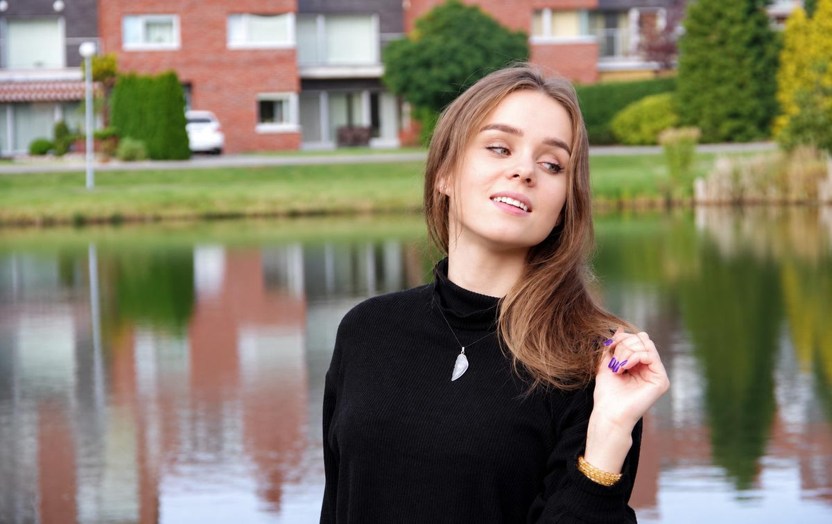 A woman wears a black turtleneck with a silver-toned wing-shaped quartz pendant necklace and a gold-coloured bracelet, standing by water.