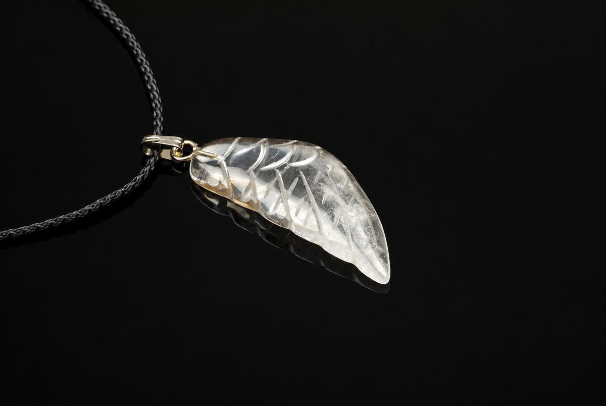 A clear quartz wing pendant has carved feather lines and a silver toned loop, hanging from a black cord against a dark background.