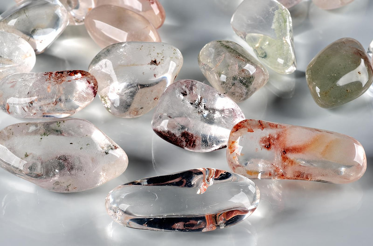 Polished quartz stones with visible inclusions, including red and green minerals, scattered on a pale surface.