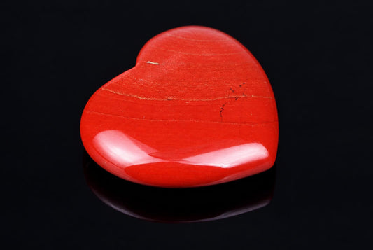 A polished red heart-shaped stone with a smooth, glossy surface and faint natural lines rests on a dark reflective surface.