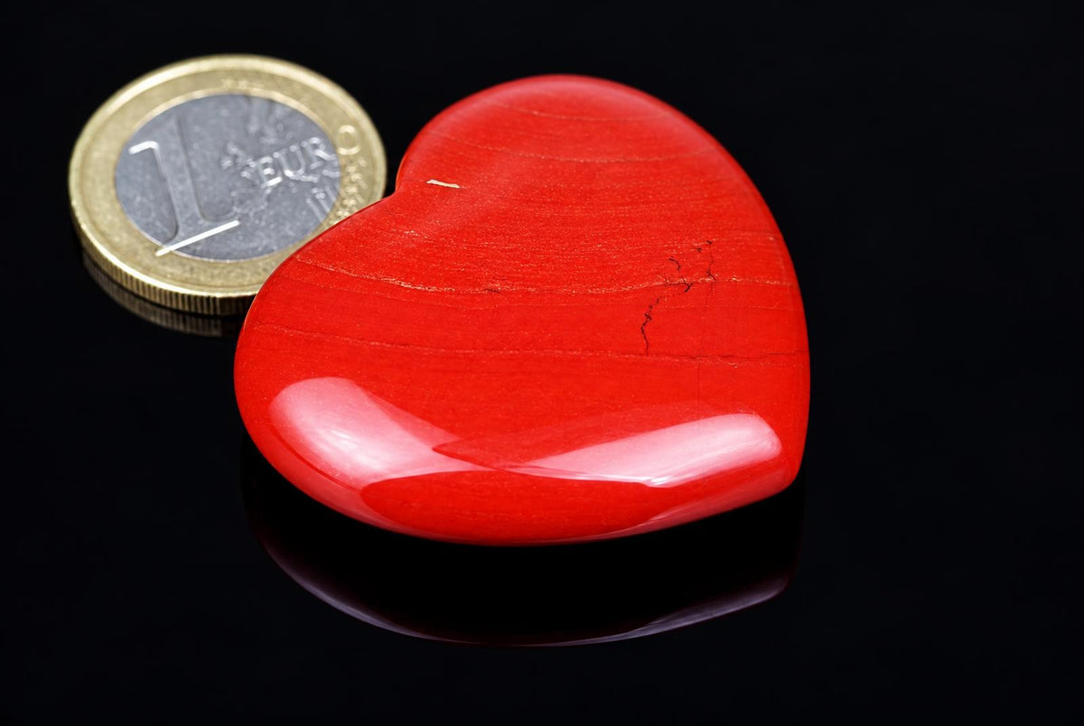 A polished red heart-shaped stone with a smooth, glossy surface sits on a dark reflective surface next to a one-euro coin.