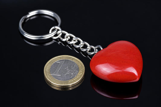 A polished red heart-shaped stone is attached to a short metal chain and keyring beside a coin on a reflective black surface.