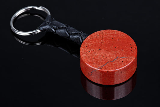 A red jasper keychain with a braided black leather strap and silver-toned metal ring, resting on a dark reflective surface.