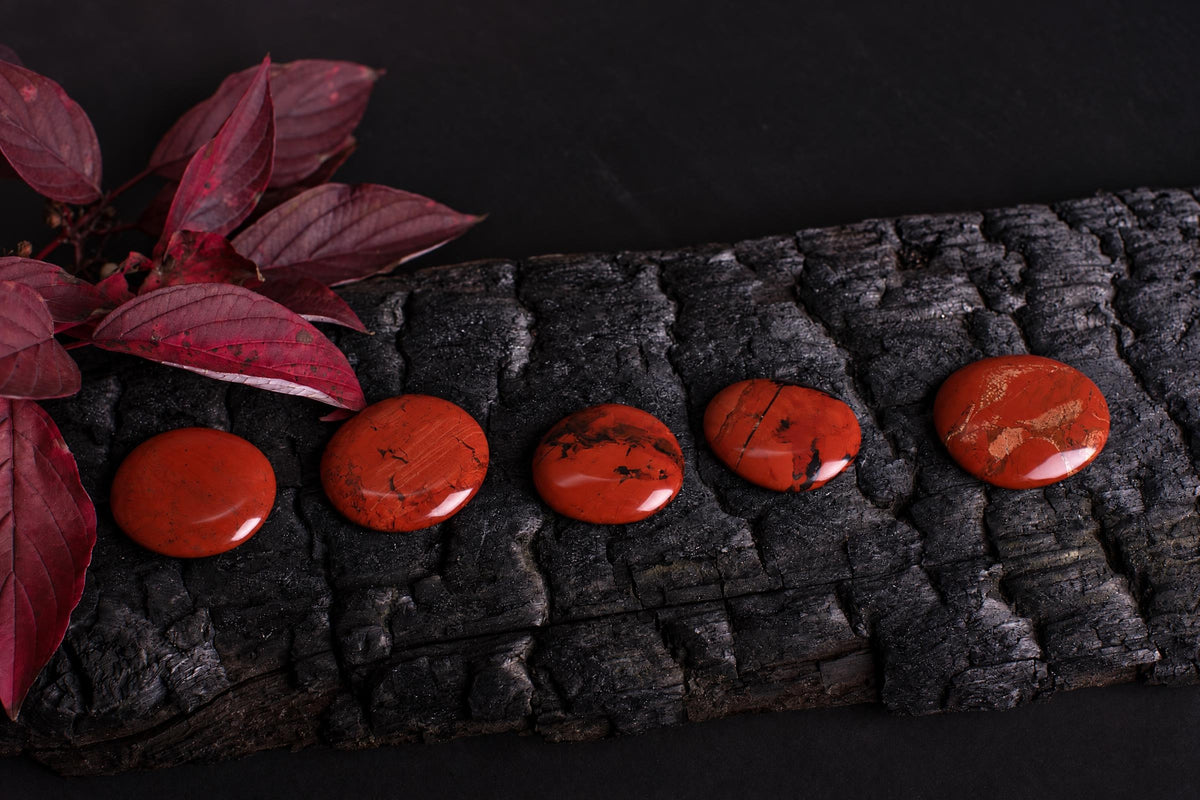Polished red jasper palmstones with dark markings are arranged in a row beside red leaves on a dark textured surface.
