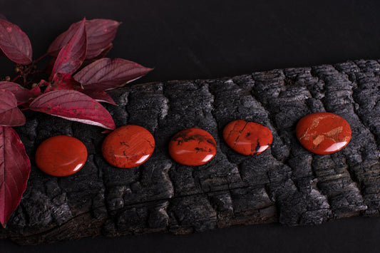 Polished red jasper palmstones with dark markings are arranged in a row beside red leaves on a dark textured surface.