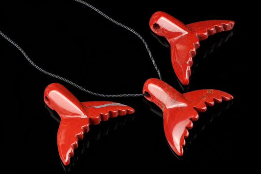 Three red jasper pendants shaped like dolphin tails hang from a twisted cord on a dark, reflective surface.