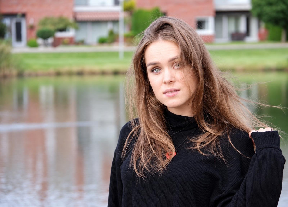 A woman with long brown hair wearing a black turtleneck sweater stands by water, holding her sleeve.