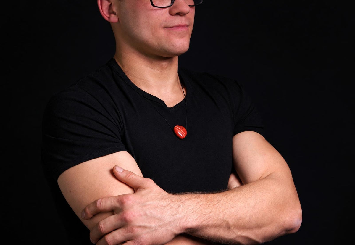 A man wears a black t-shirt with a red jasper pendant shaped like a heart on a dark surface.