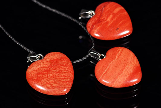 Three red jasper heart pendants on silver-toned cords, resting on a dark reflective surface.
