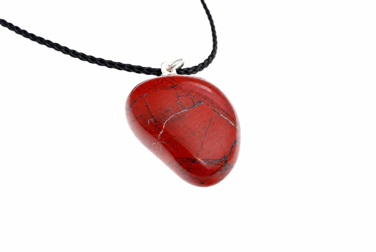 A smooth red jasper pebble pendant shows thin dark veins and a silver-colored loop, hanging from a black cord on white.