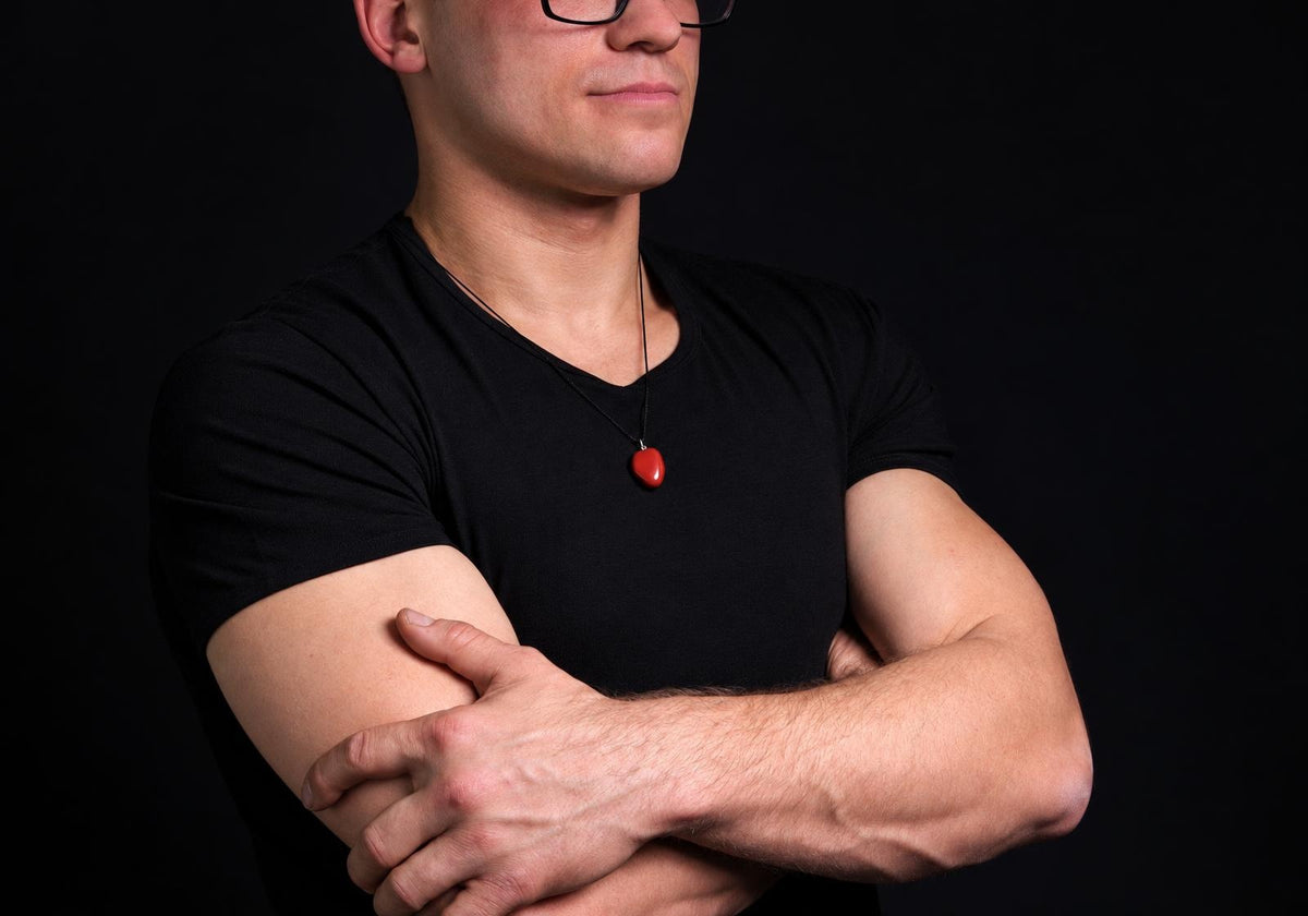 A man wears a black t-shirt and glasses, with arms crossed against a dark background. He has a necklace with a small red heart-shaped pendant.