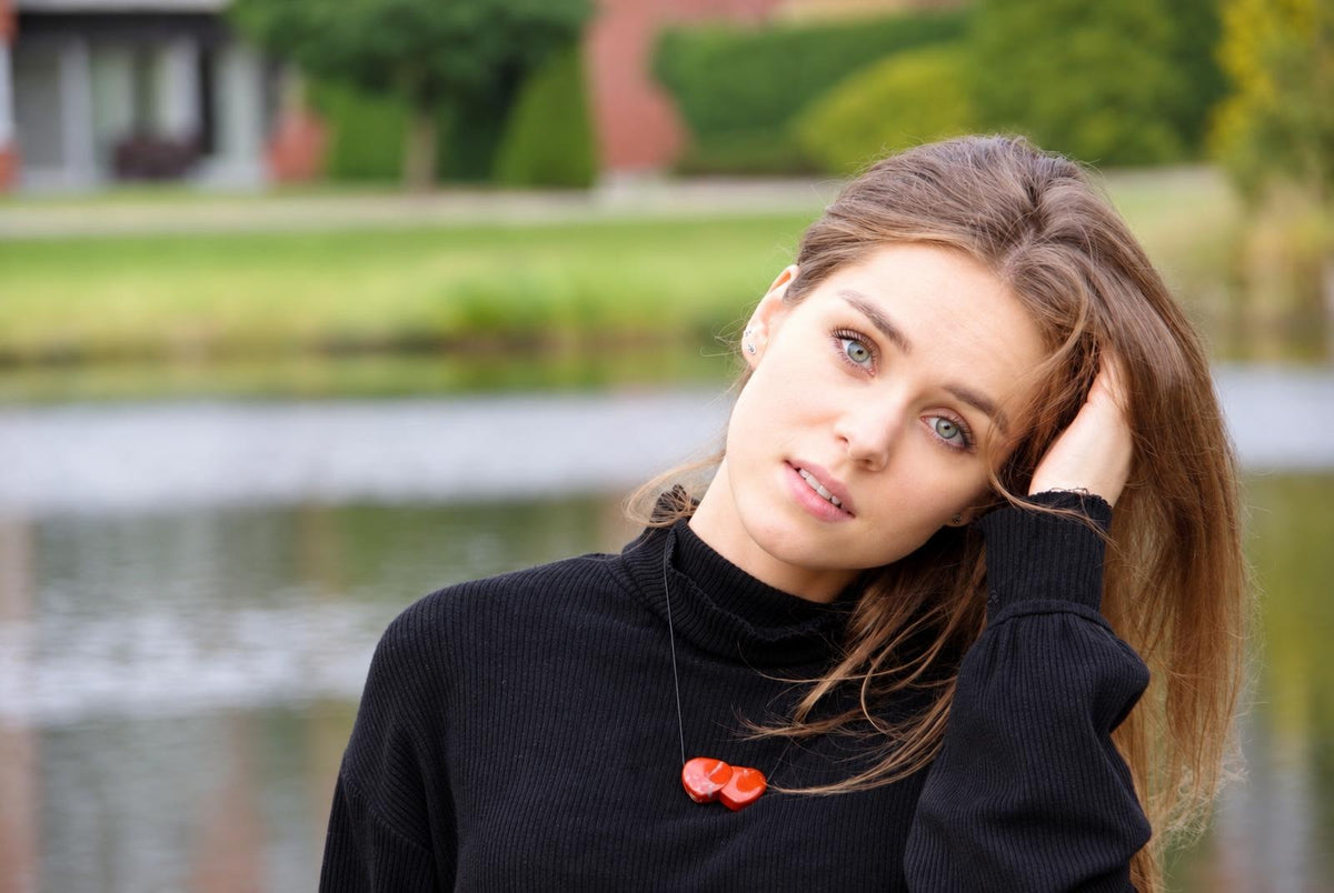 A woman wears a black turtleneck with a red pendant necklace shaped like two joined hearts, set against a blurred outdoor background.