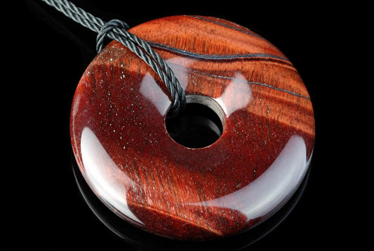 A polished, donut-shaped pendant with red and brown wood grain patterns, a central hole, and a grey cord looped through it against a dark surface.