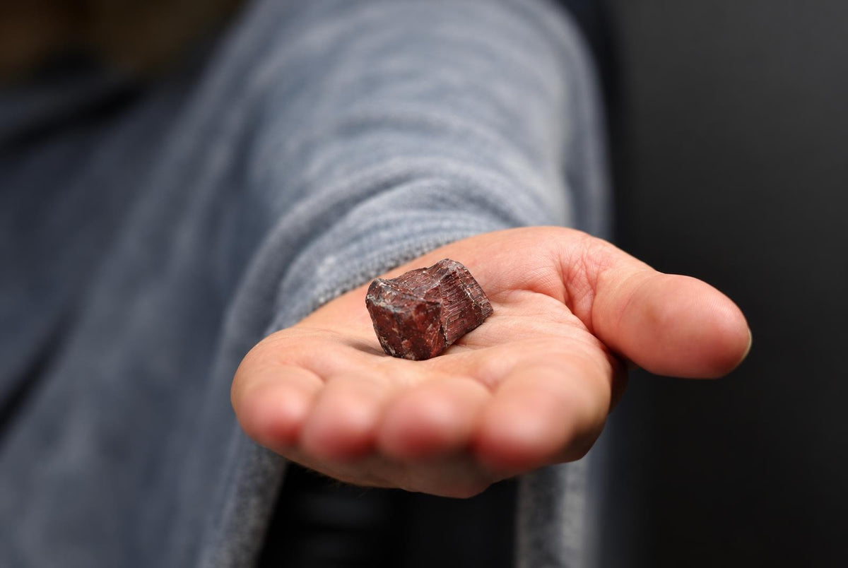 A person holds a small, reddish-brown raw stone with visible striations in their open palm against a dark background.