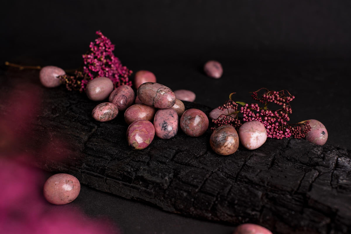 Polished pink rhodonite stones with black and white patterns rest on a dark wooden surface, accompanied by sprigs of small pink flowers.
