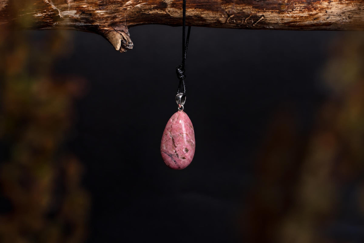 A pink rhodonite pendant with a silver bail hangs from a black cord on a wooden branch against a dark background.