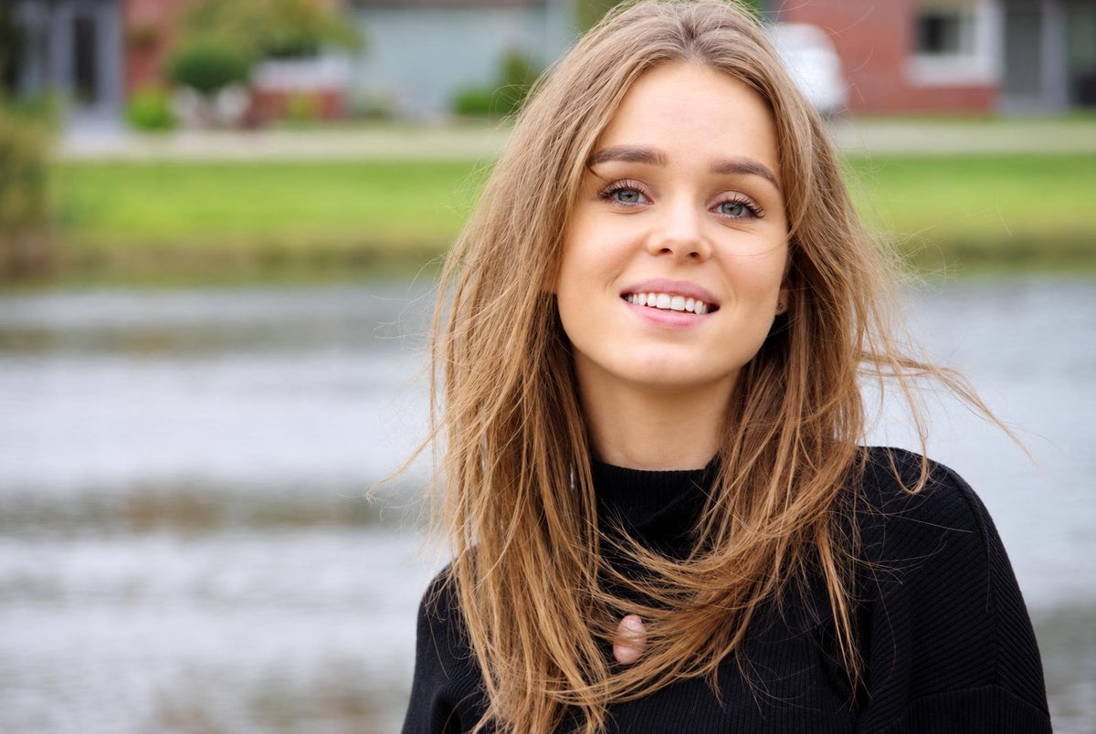 A smiling woman with long light brown hair wearing a black top, outdoors near water.