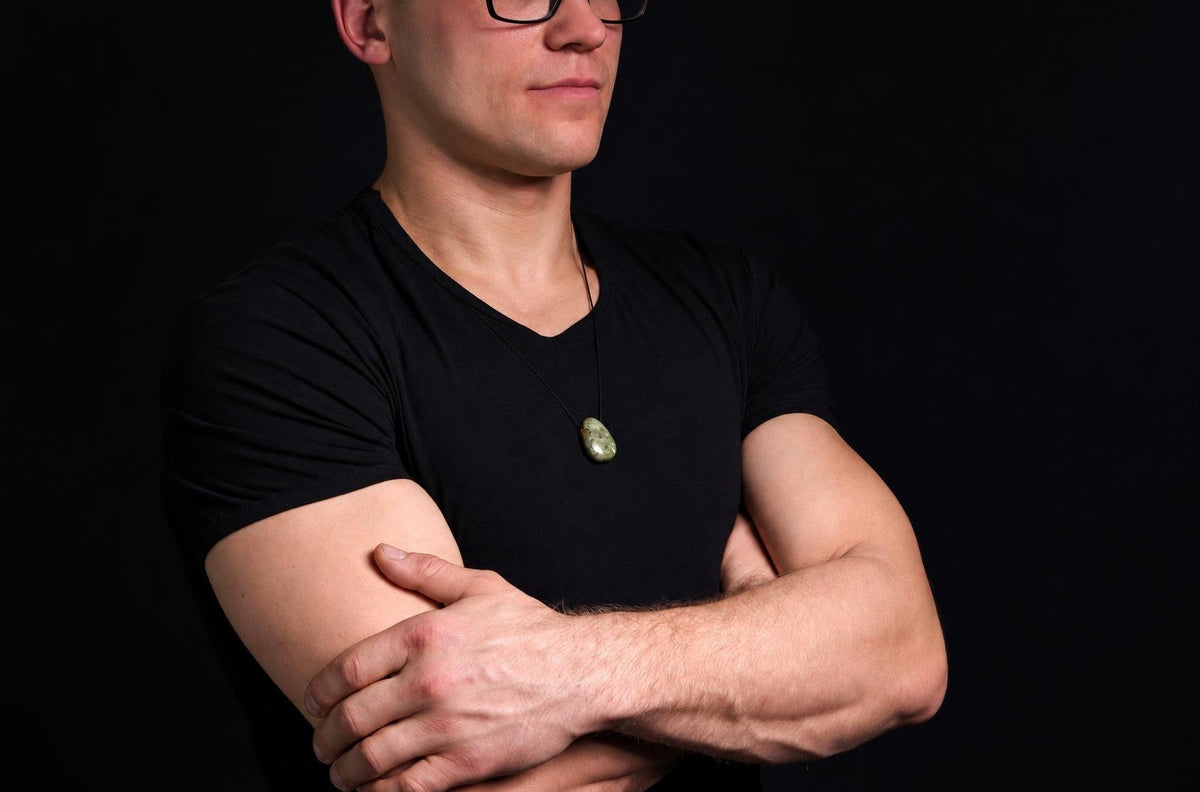 A man in a black t-shirt wears a green, oval-shaped pendant on a dark cord against a dark background.