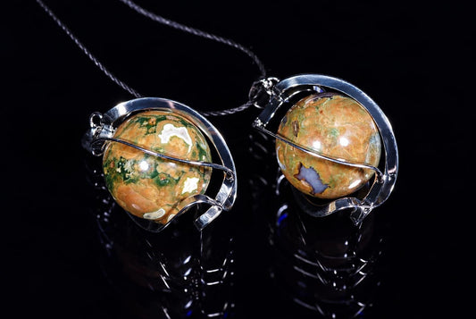 Two silver-toned pendants with round, polished stones in green, orange and white patterns on a dark reflective surface.