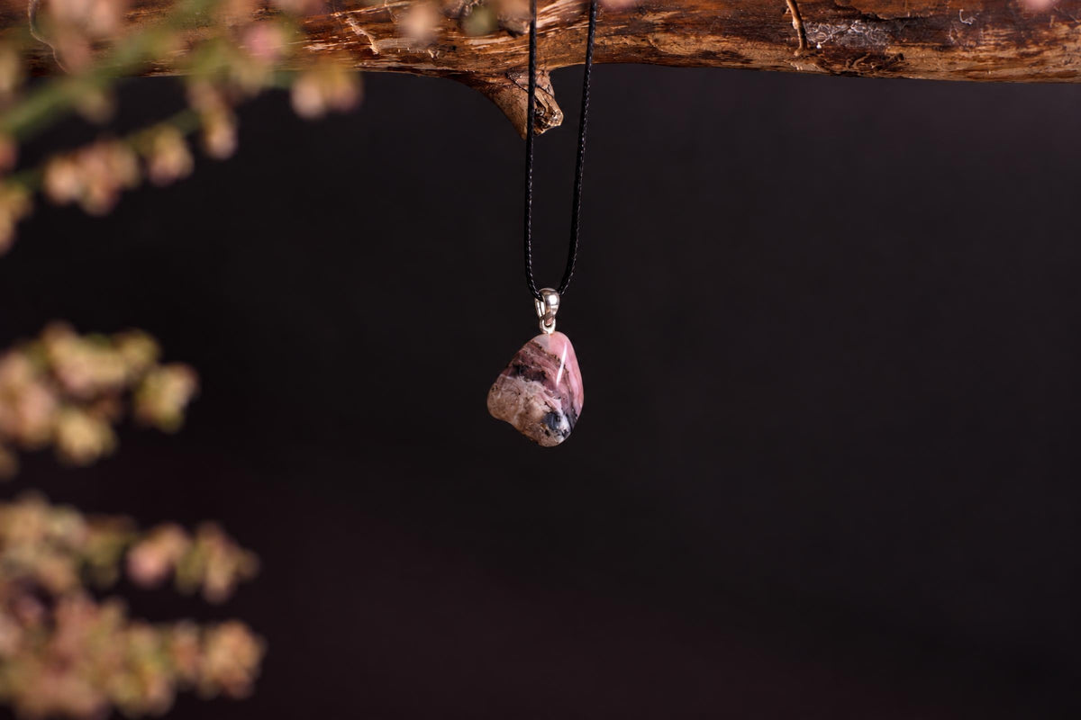 A pendant with a pink, brown and black polished stone hangs from a black cord on a dark surface.