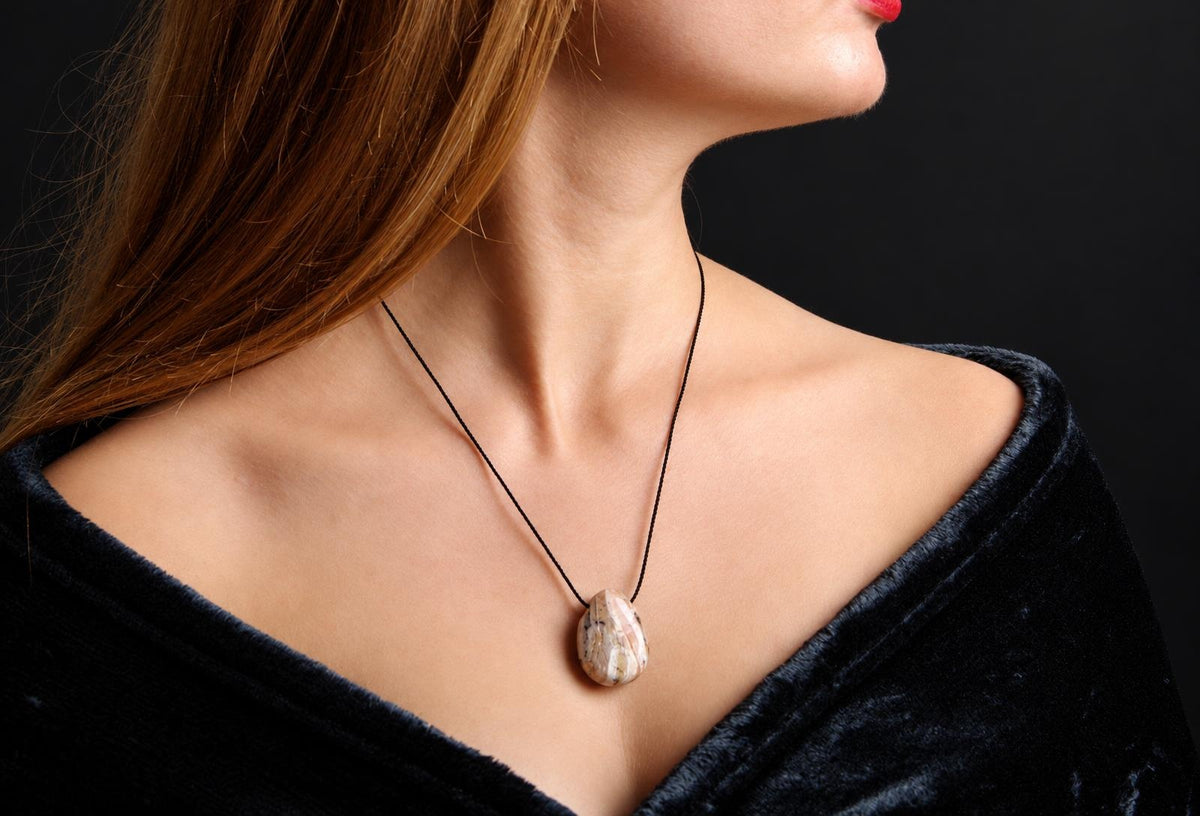 A woman wears a pendant necklace with an oval, cream-coloured stone featuring dark speckles on a black cord against a dark surface.