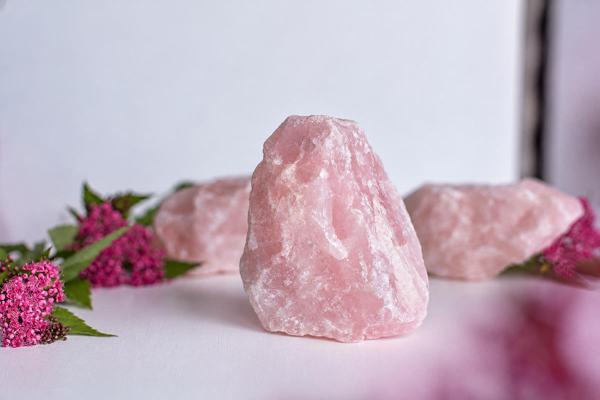 Three pink rose quartz crystals on a pale surface with pink flowers nearby.